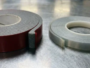 Acrylschaum-Klebebandrolle und dünnes doppelseitiges Trägerband nebeneinander auf Stahlwerkbank, Querschnitt zeigt Schaum- und Klebeschichten.